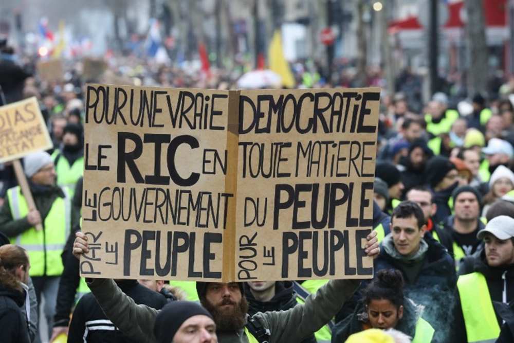 Un manifestant avec un gilet Jaune tient une pancarte ou est inscrit, pour une véritable démocratie le ric en toute matière, le gouvernement du peuple, par le peuple, pour le peuple