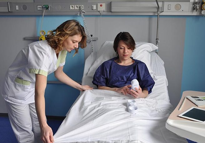 Une patiente dans une chambre individuelle, avec une infirmière qui la borde, elle a accès au téléphone, elle a une tablette informatique sur la table devant elle